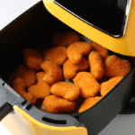 Fritadeira elétrica amarela com o cesto aberto cheio de nuggets dourados, ilustrando qual a potência ideal para uma boa air fryer cozinhar alimentos congelados com eficiência.